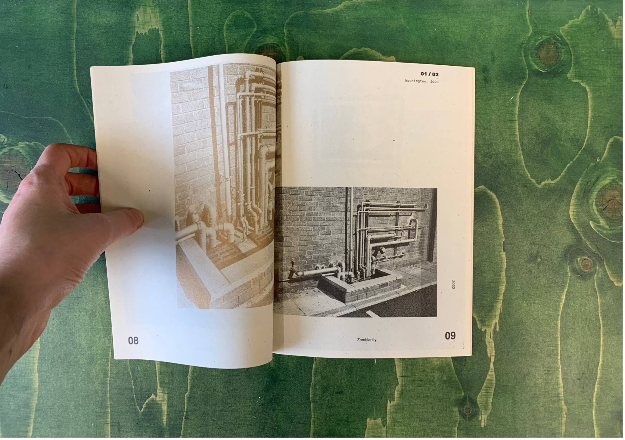 A photograph of a hand holding open a book on a green table. The books pages are white. There are two images printed across either page showing a large number of pipes going into the ground. The image on the left is printed in gold ink and the image on the right is printed in black and white.