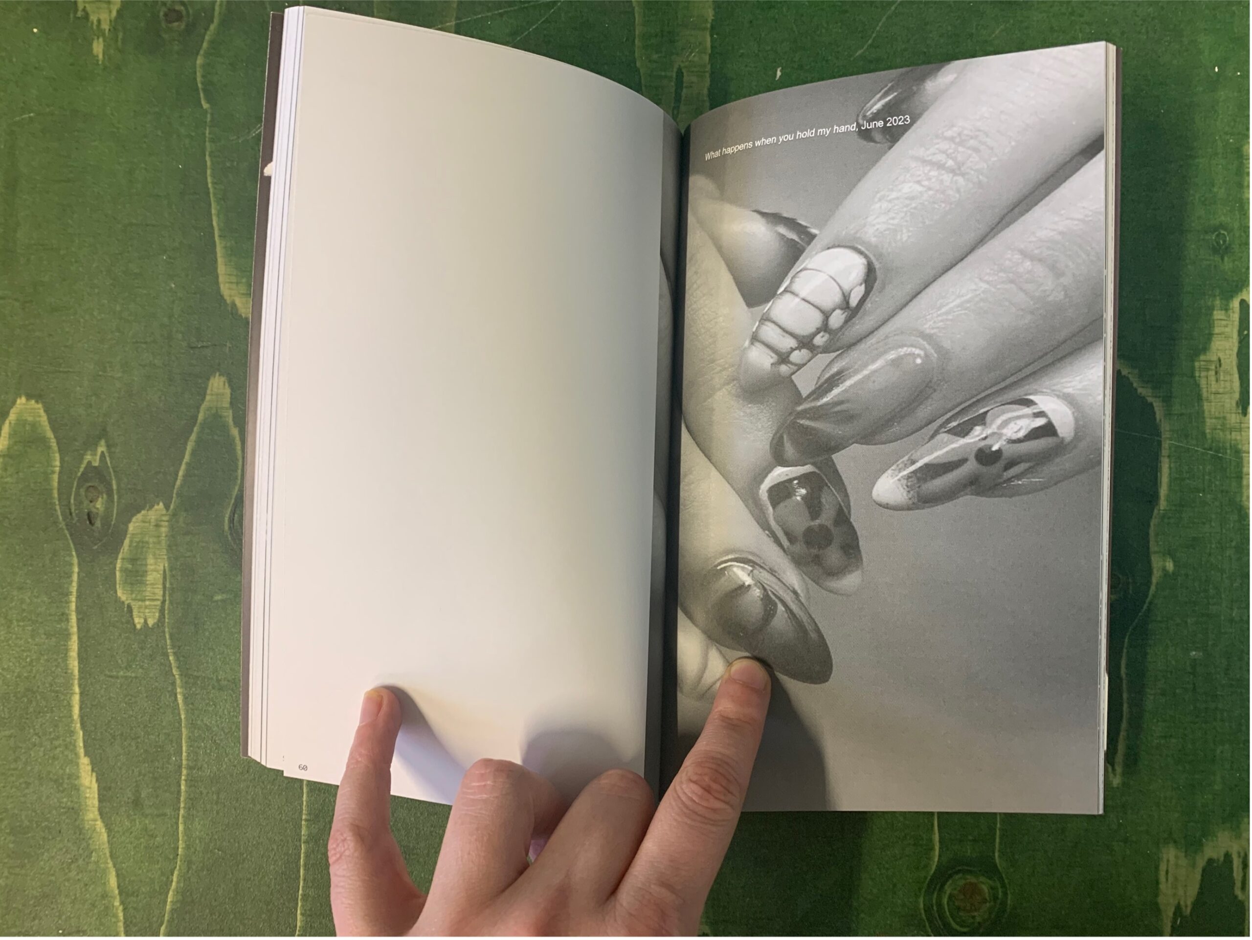 A photograph of a hand holding open a book on a green table. The left page is blank and the right page shows a black and white photograph of a pair of hands with false nails on.